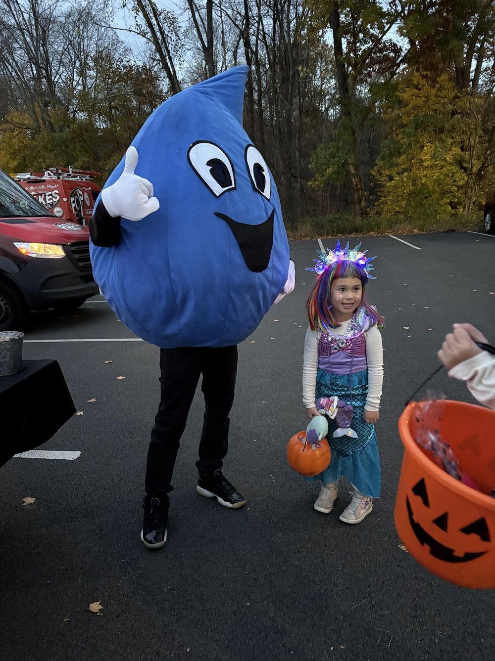 A person in a blue water drop mascot costume gives a thumbs-up next to a child wearing a lit tiara and mermaid costume.