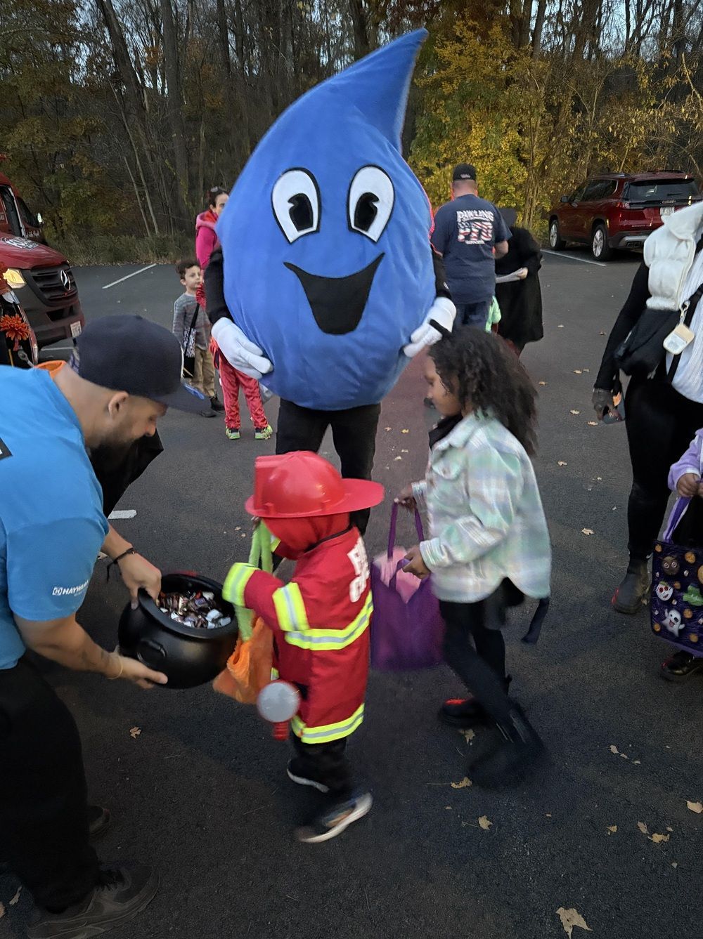 A person in a blue water drop mascot costume hands candy to a child dressed as a firefighter at an outdoor event.
