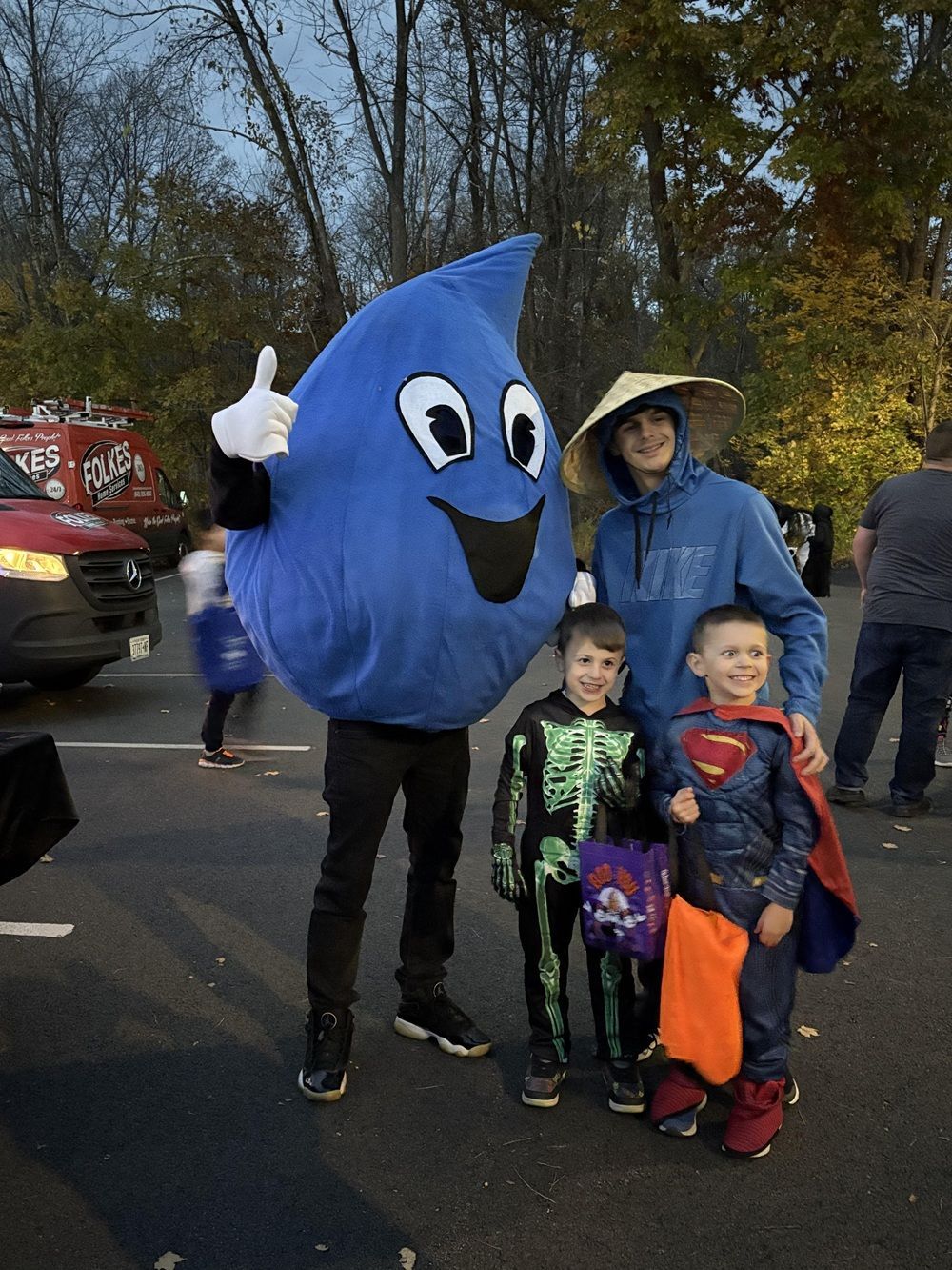A mascot in a blue water drop costume poses with three people dressed in Halloween costumes at an outdoor event.