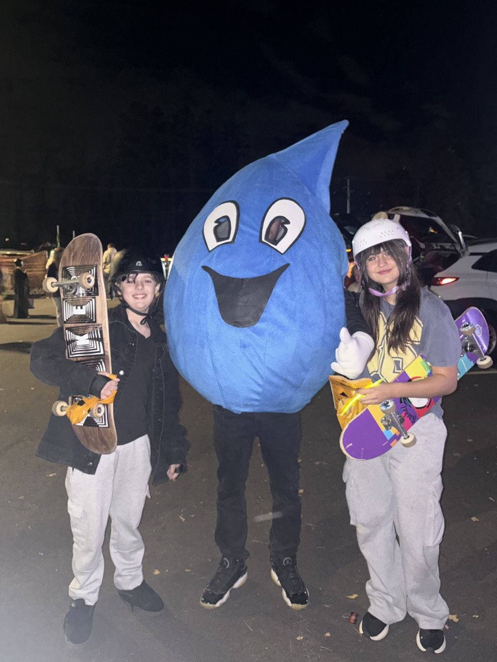 Two people with skateboards stand on either side of a large blue water drop costume mascot at night.