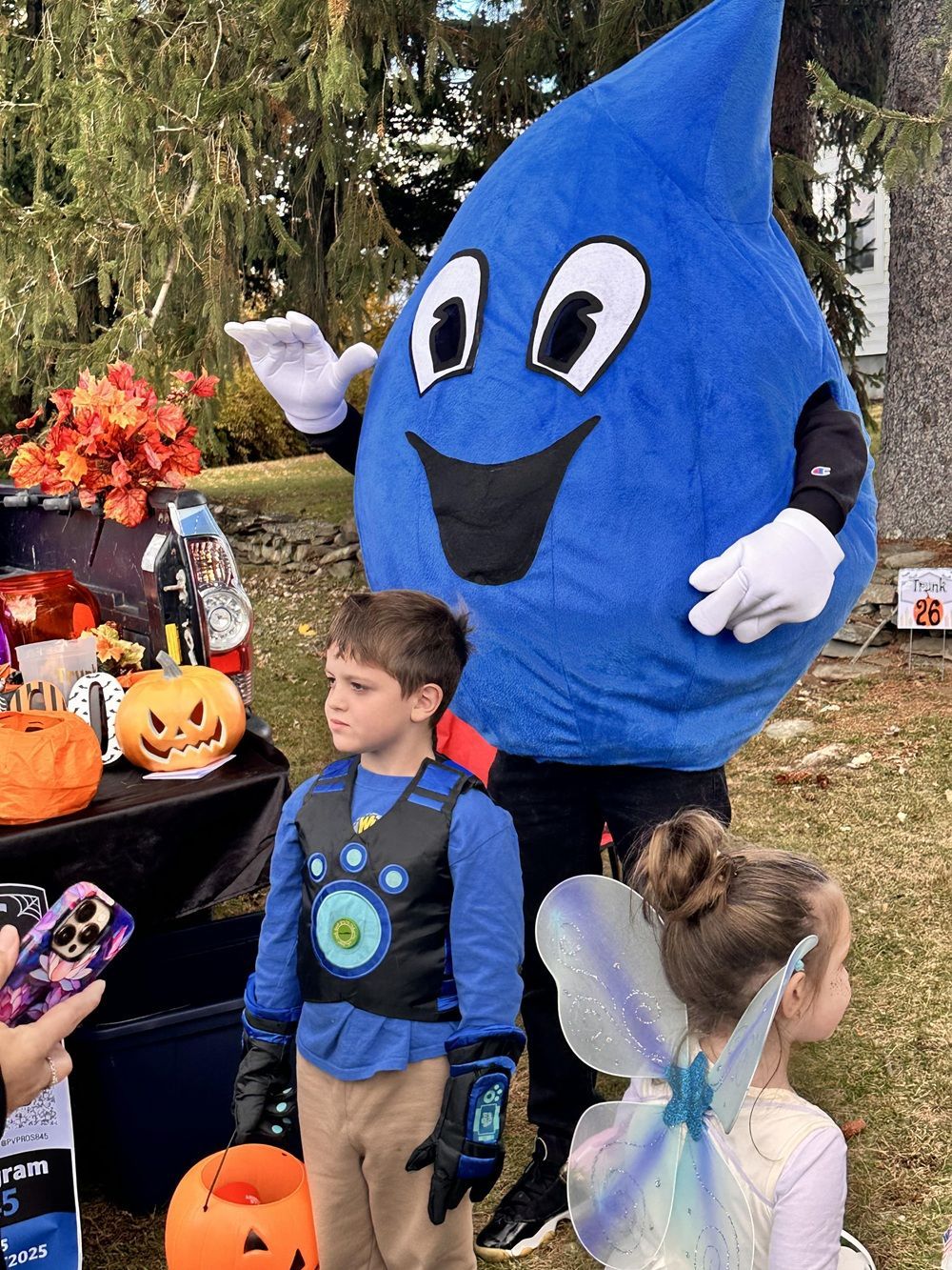 A large blue water-drop mascot waves next to two children in Halloween costumes outdoors near a pumpkin display.