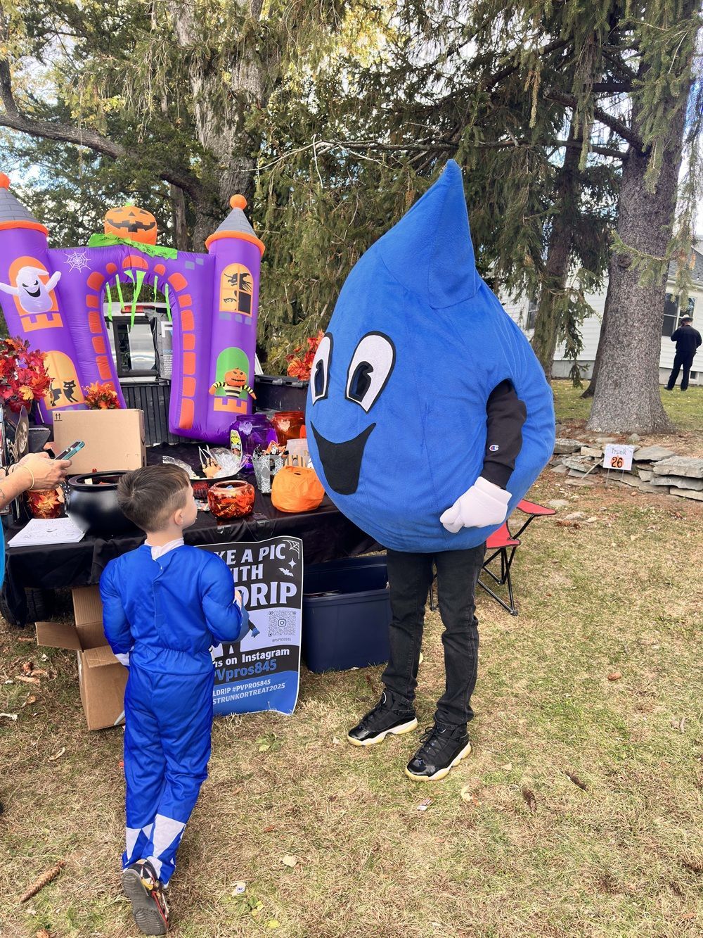 A child in a blue costume faces a person in a large blue water drop mascot suit at an outdoor event with decorations.