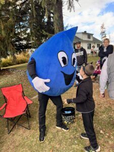 A person in a blue water droplet mascot costume interacts with a child outdoors on a grassy area near a house.