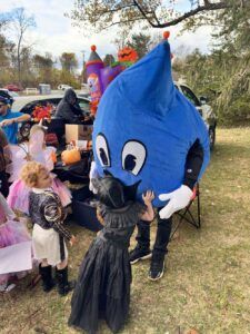 A child dressed as Wednesday Addams hugs a blue, teardrop-shaped mascot at an outdoor Halloween event.