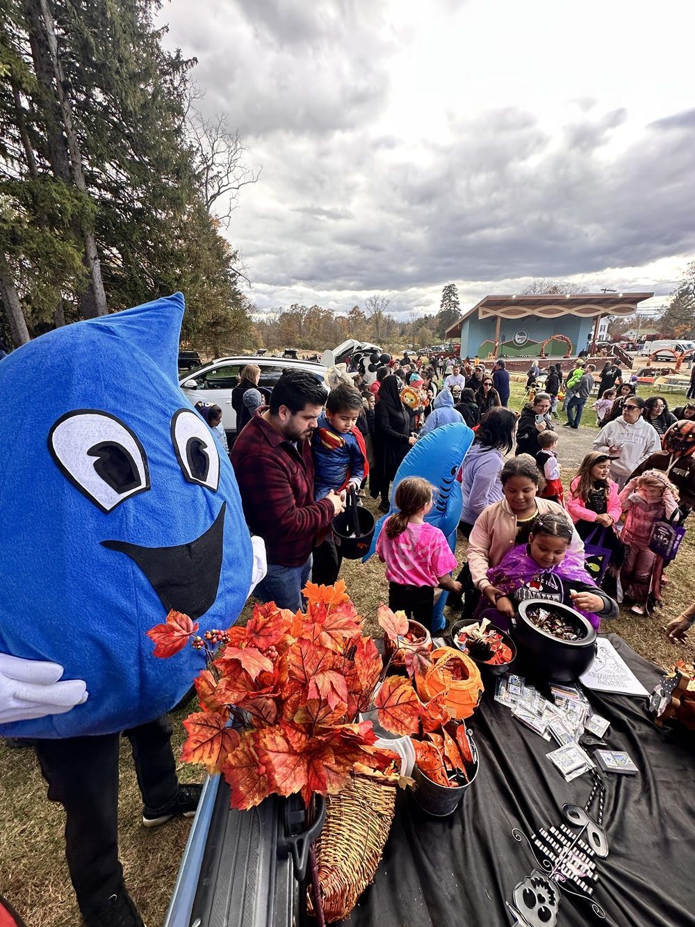 A blue raindrop mascot stands near a table decorated with orange autumn leaves at an outdoor fall event.