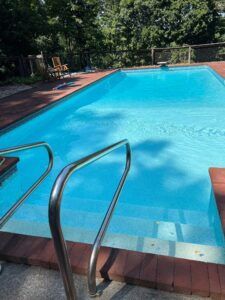 A rectangular swimming pool with bright blue water and metal handrails, set in a backyard with trees in the background.