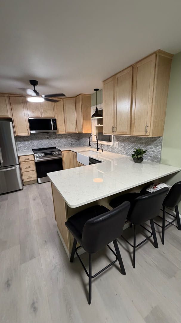A modern, U-shaped kitchen featuring light wood cabinets, white countertops, stainless steel appliances, and bar stools.
