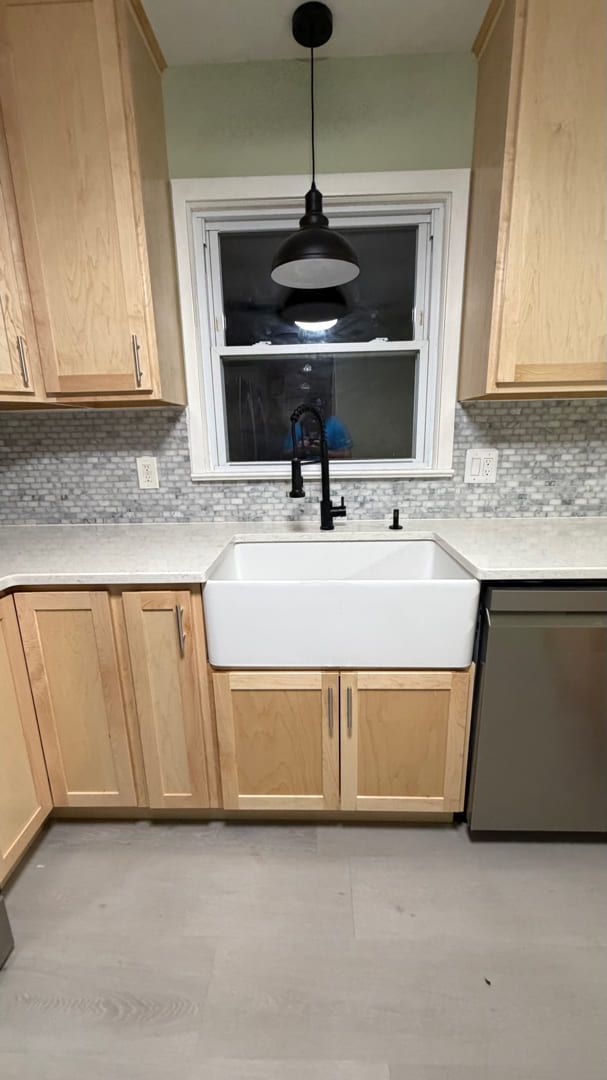 Kitchen view with light wood cabinets, white farmhouse sink, black faucet, grey tile backsplash, and hanging pendant lamp.