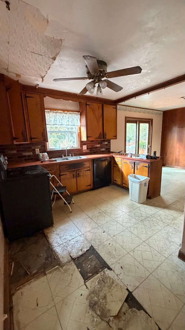A dated, fixer-upper kitchen featuring wood cabinets, a ceiling fan, and damaged, partially missing floor tiles.