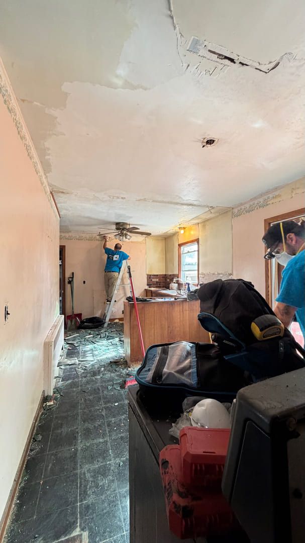 Two people in a home interior under construction, with a damaged, peeling ceiling and debris scattered on the floor.