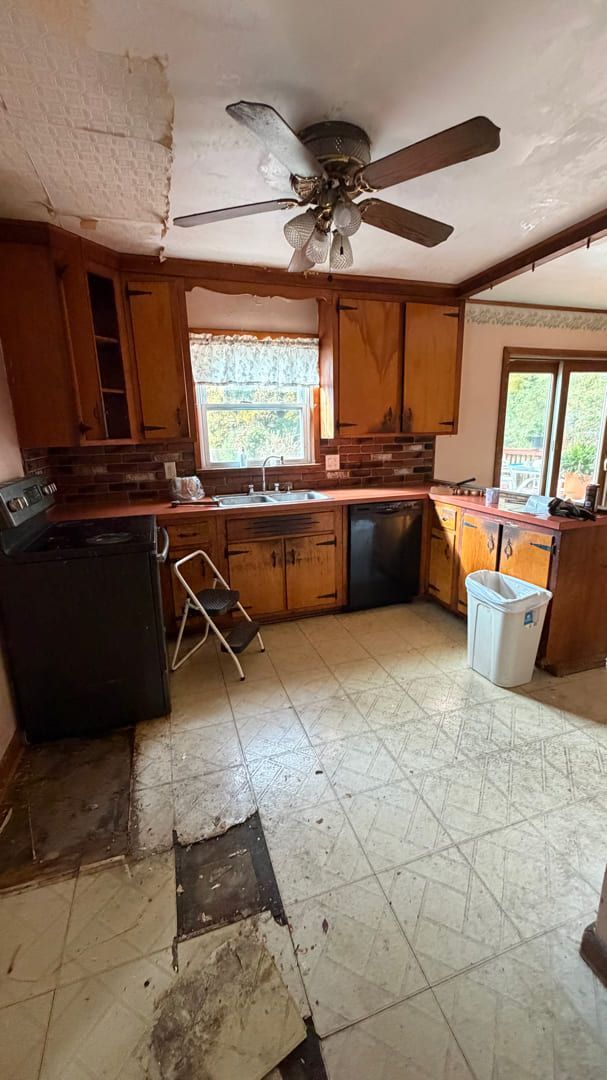 A kitchen with worn wood cabinets, black appliances, a ceiling fan, and damaged vinyl flooring.