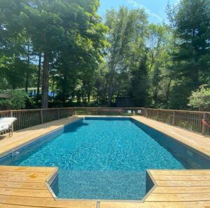 A rectangular backyard swimming pool surrounded by a wooden deck and a forest of tall green trees on a sunny day.