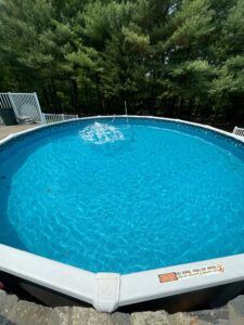 A round above-ground swimming pool with clear blue water and a small fountain, set against a background of green trees.