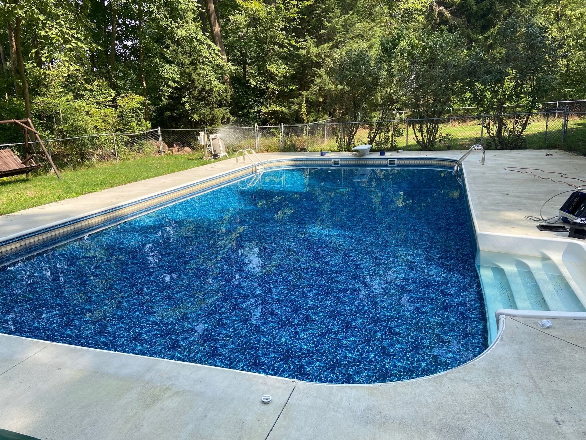 A backyard swimming pool with a deep blue tiled pattern and white steps, surrounded by a concrete deck and trees.