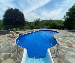 A blue, kidney-shaped in-ground swimming pool surrounded by a stone patio on a sunny day with trees in the background.