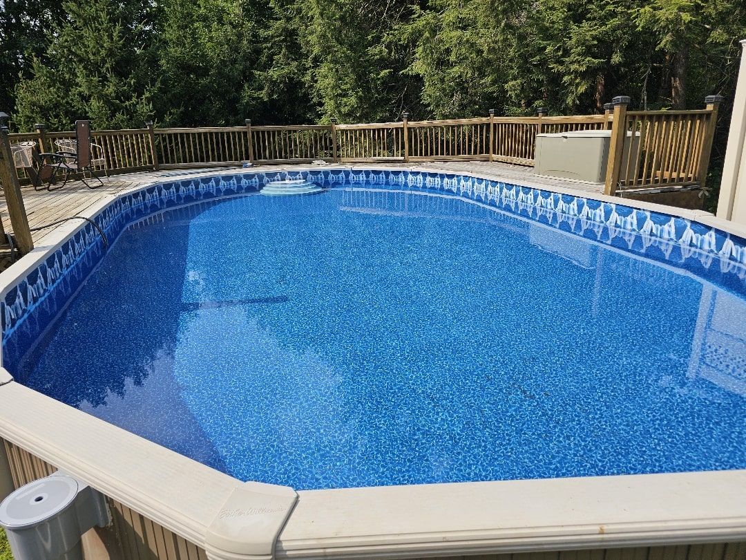 An oval above-ground swimming pool filled with blue water, surrounded by a wooden deck and lush green trees.