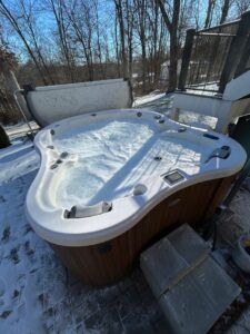 A triangular hot tub with bubbling water and a lid lifted open, set on a snowy deck outside near trees.