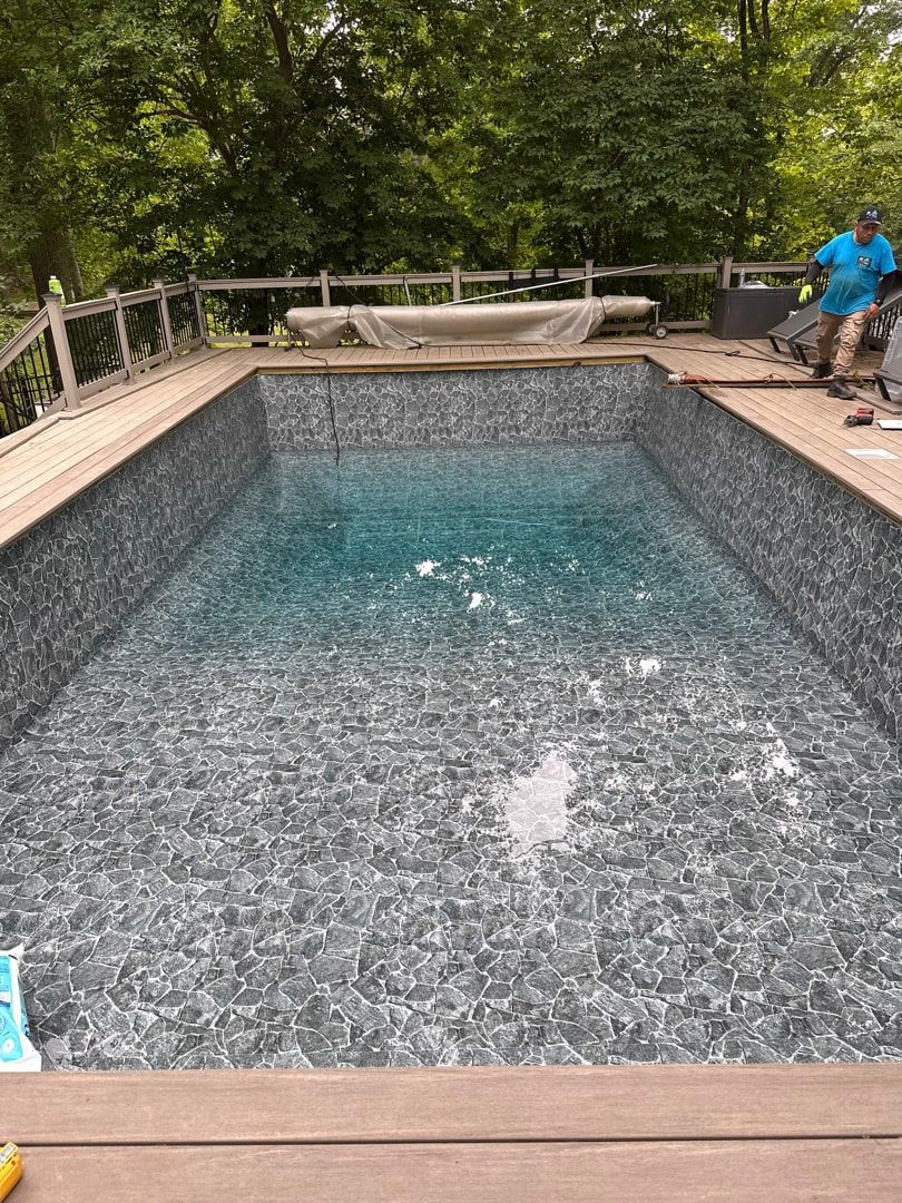 A rectangular swimming pool with a patterned dark grey liner sits on a deck, with a person standing near the edge.