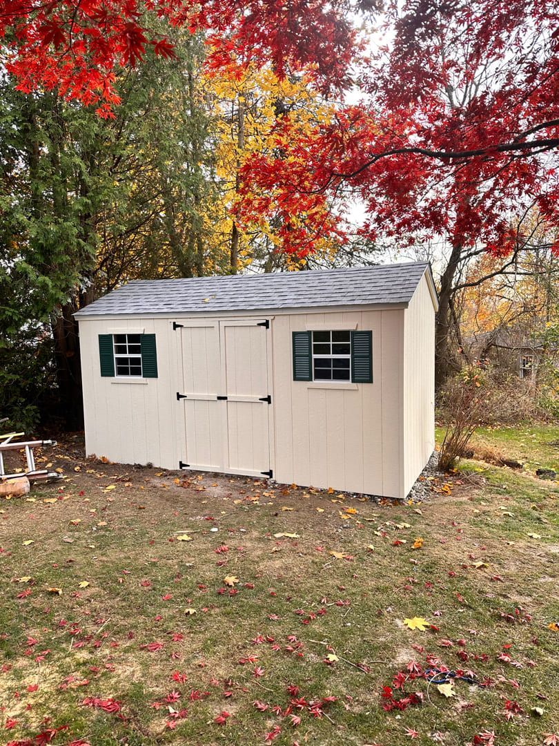 A small, off-white wooden shed with green-shuttered windows stands on a lawn surrounded by colorful autumn trees.
