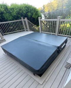 A square hot tub with a dark grey cover sits on a light-colored wooden deck surrounded by a railing and green trees.