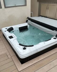 A white hot tub with a black base and a partially open grey cover, set into a wooden deck next to a building wall.