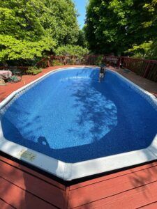 An oval swimming pool with a blue liner surrounded by a reddish-brown wooden deck and green trees.