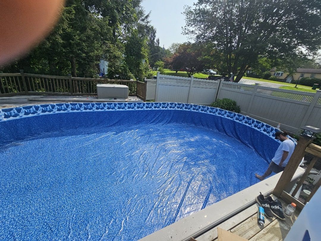 A person stands on a wooden deck next to an oval-shaped above-ground swimming pool with a blue-tiled pattern liner.