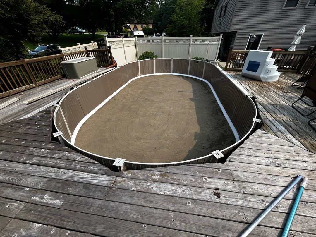 An empty, oval-shaped above-ground swimming pool shell with a dirt floor, surrounded by a wooden deck.