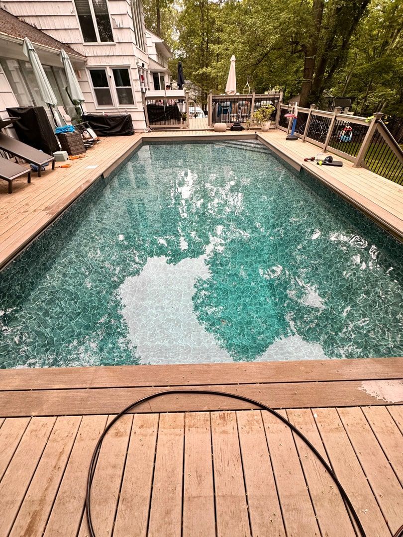 A rectangular swimming pool with teal water sits centered in a wooden deck next to a house, surrounded by trees.
