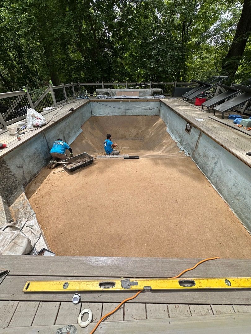Two workers in blue shirts level sand in the deep end of an empty backyard swimming pool during a renovation project.