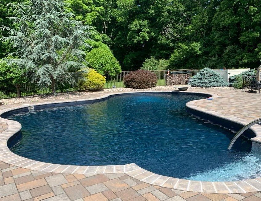 A curved swimming pool with dark blue water, surrounded by light-colored stone pavers and lush green trees in the background.