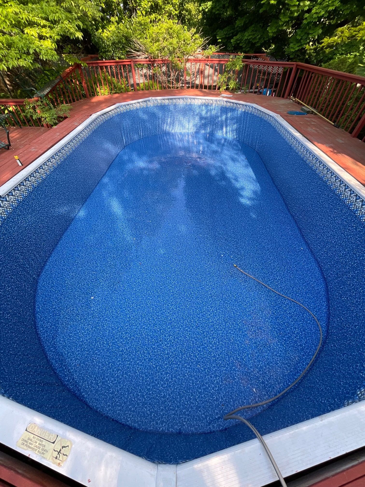 A bright blue, oval-shaped above-ground pool surrounded by a wooden deck, with lush green trees in the background.