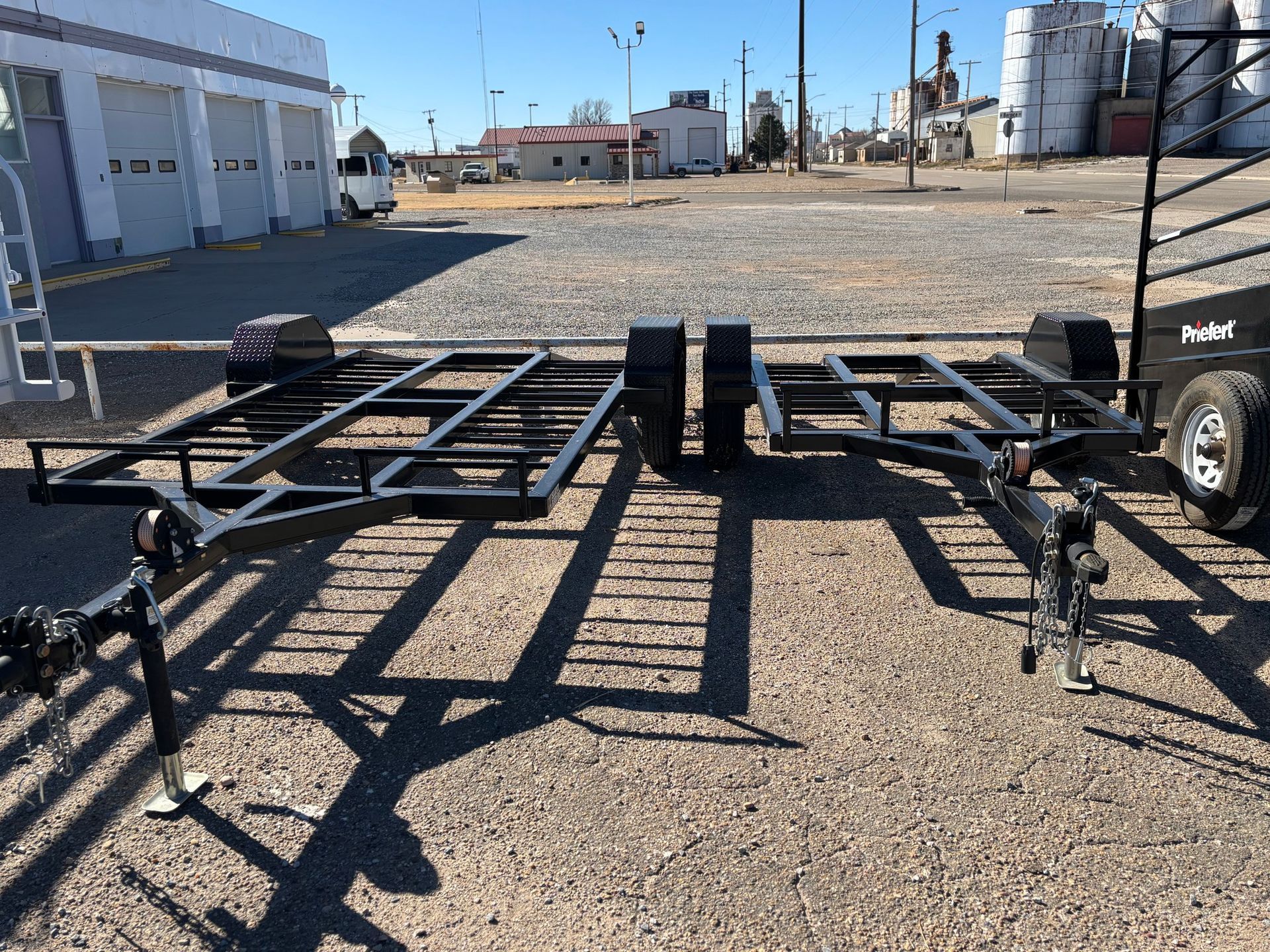 Two empty black metal trailer frames parked on a gravel lot in front of a white warehouse building on a sunny day.