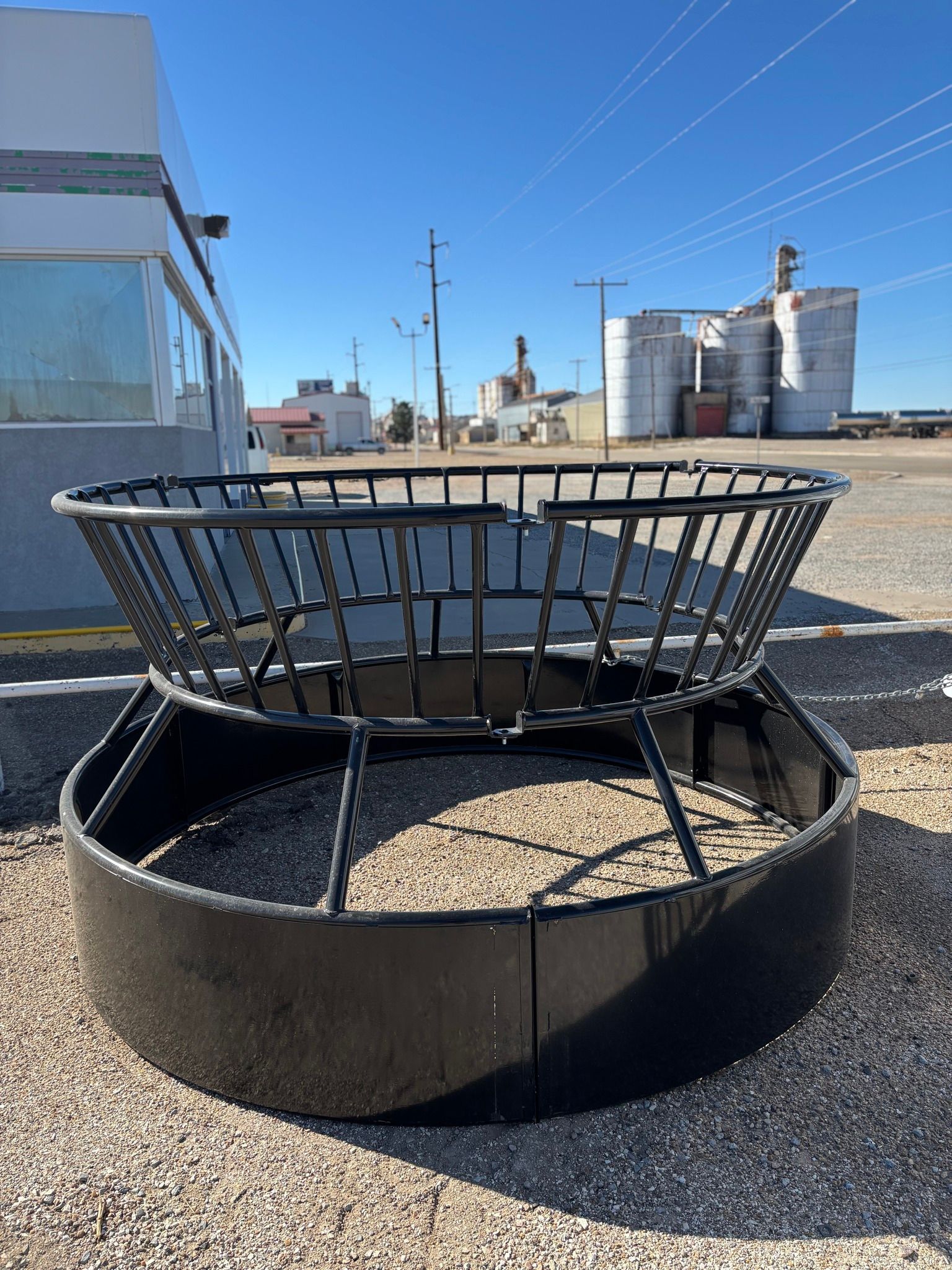 A round black metal livestock hay feeder sits on a gravel lot in front of industrial silos under a clear blue sky.