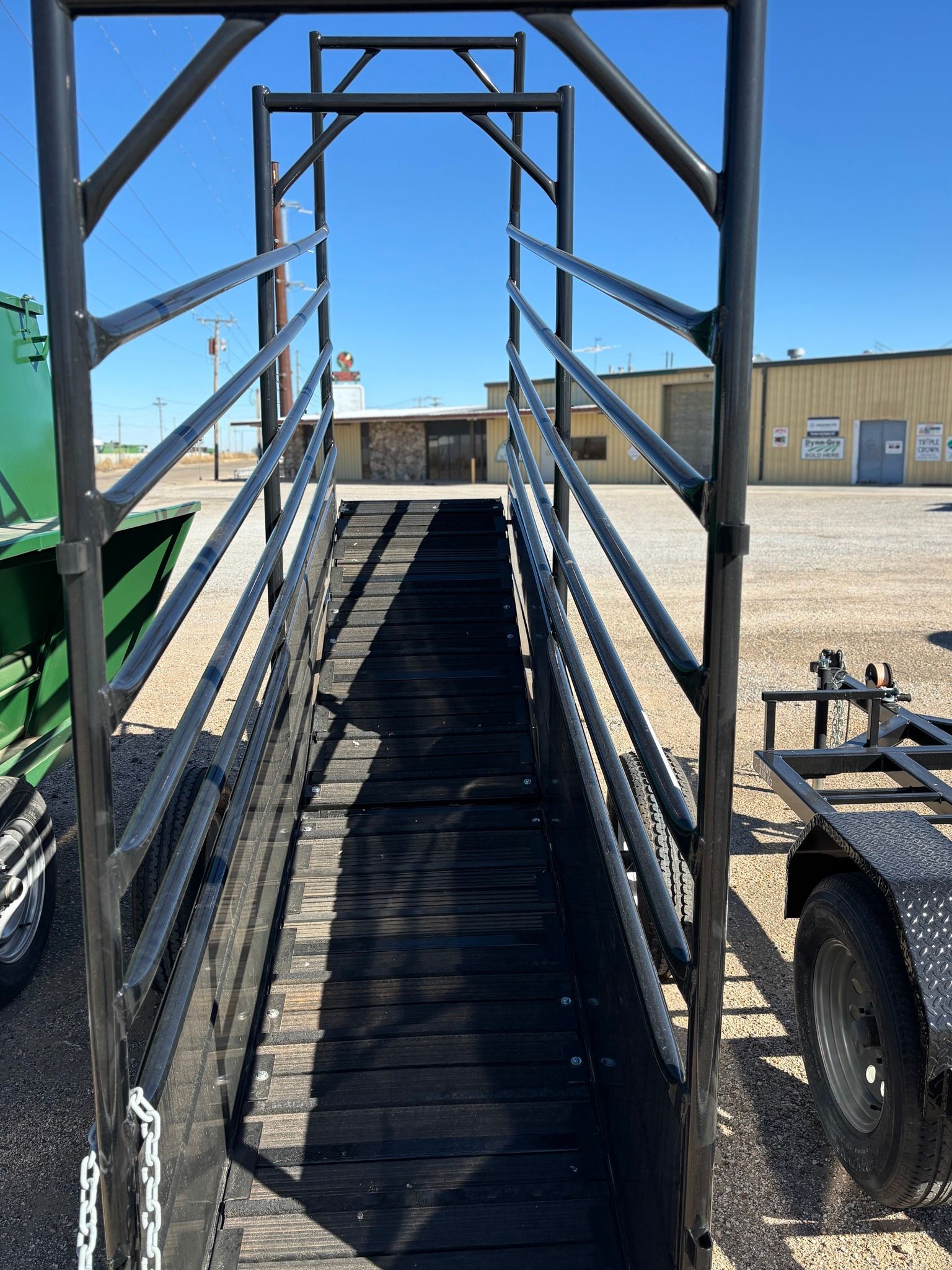 A metal livestock loading ramp with black railings and a wooden floor, set outdoors on a gravel lot under a clear sky.