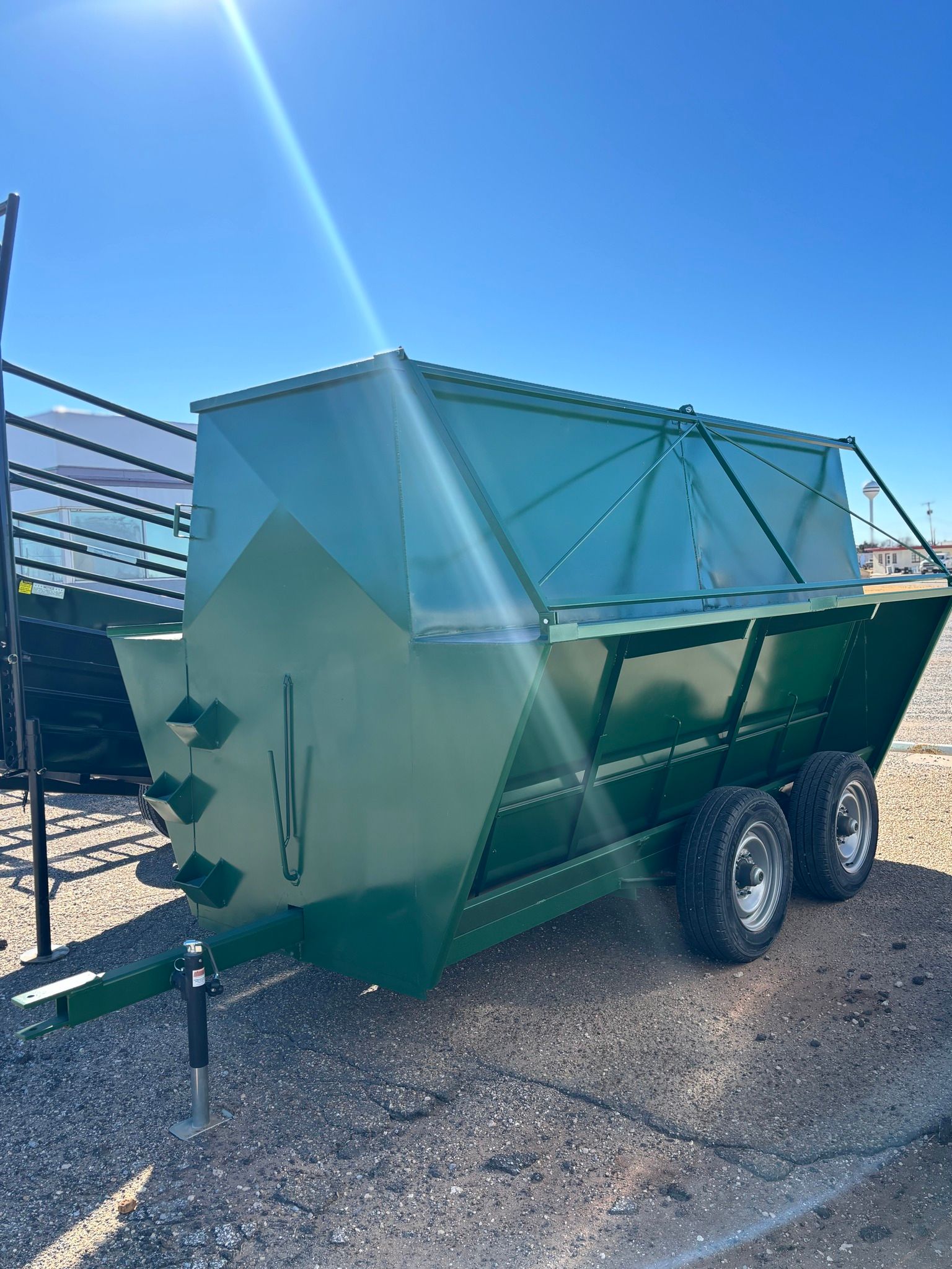 A green, tandem-axle agricultural feeder trailer parked on a gravel lot under a clear blue sky.