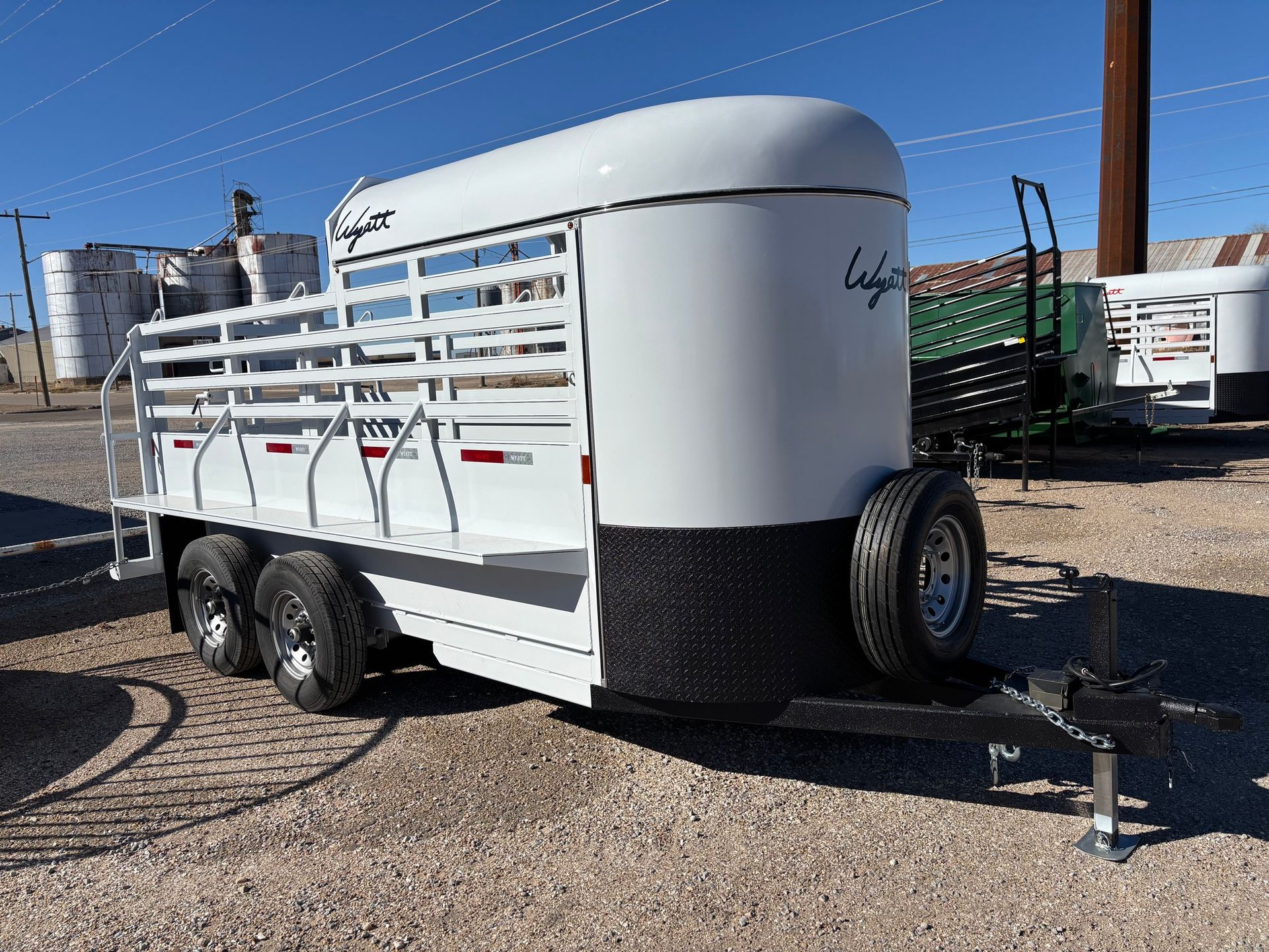 White livestock trailer with a black diamond-plate front and spare tire, parked on a gravel lot under a clear blue sky.