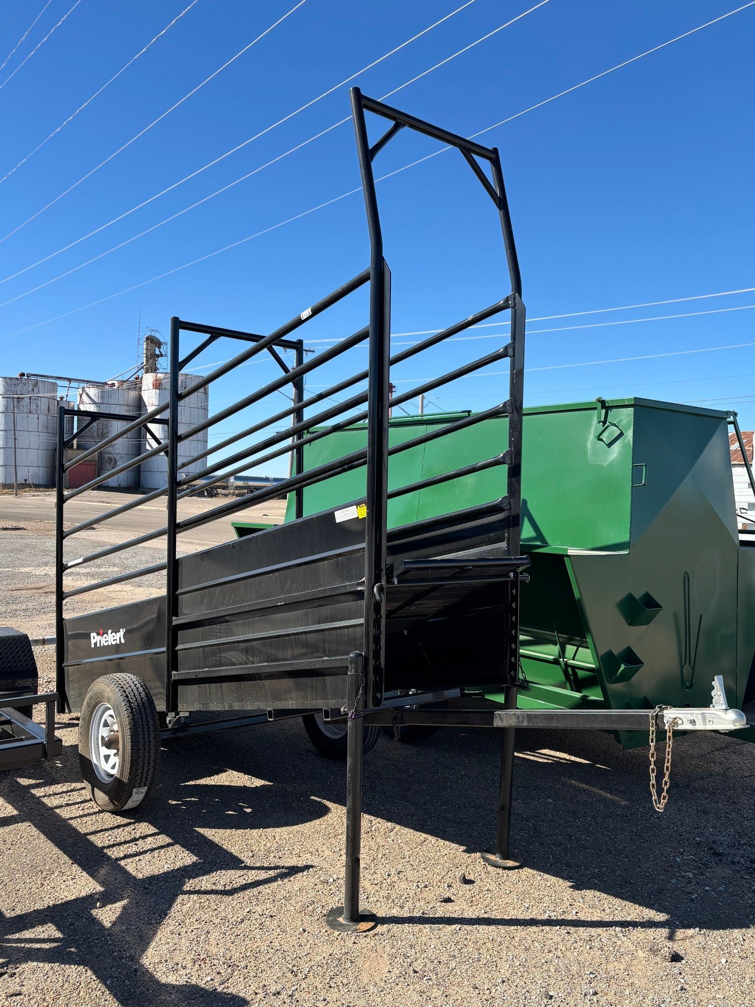 A black metal livestock trailer with high, slatted side rails and a tall frame, parked on a gravel lot under a blue sky.