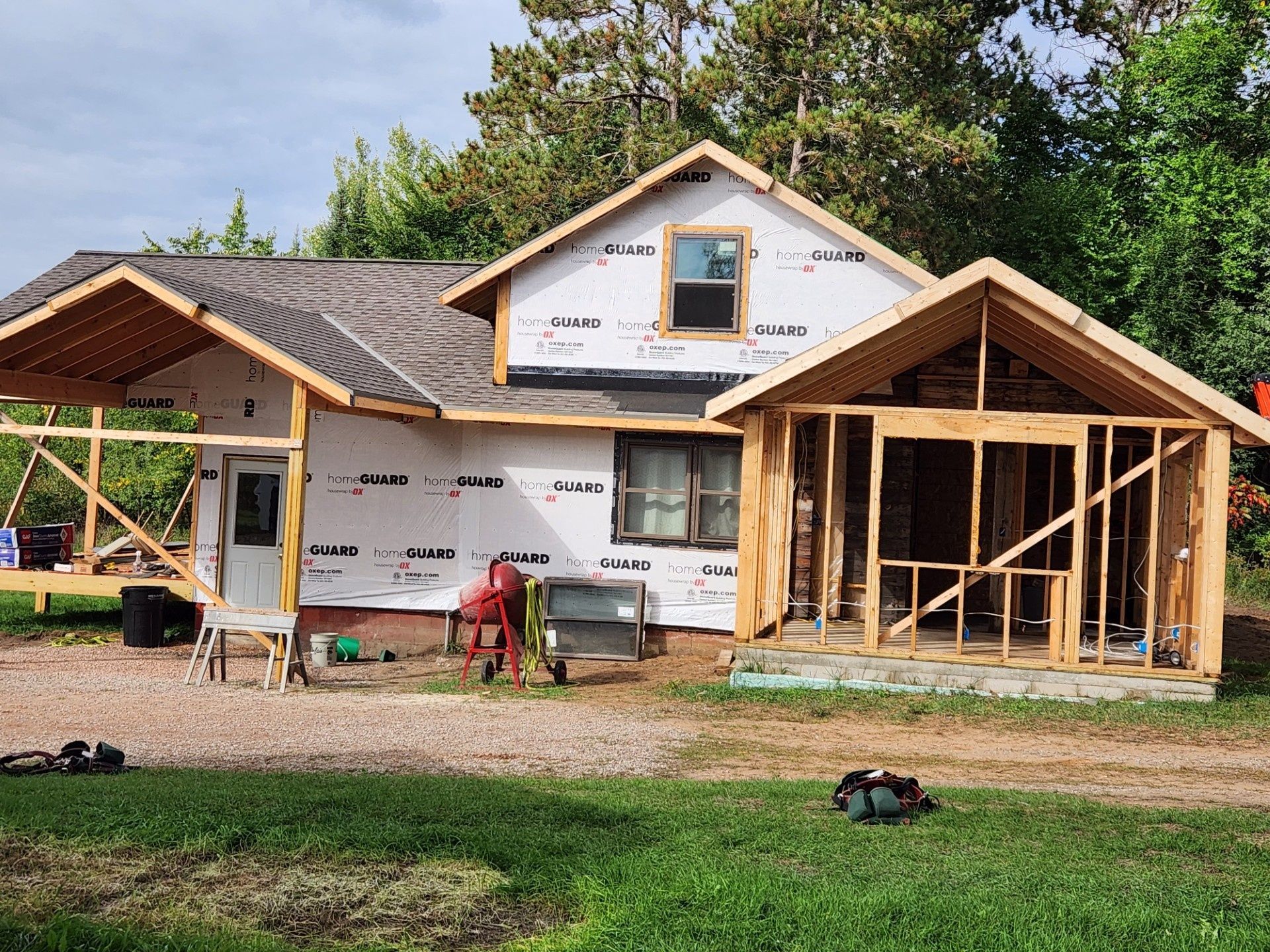 House under construction with exposed framing, wrap sheathing, and a dirt yard surrounded by trees.