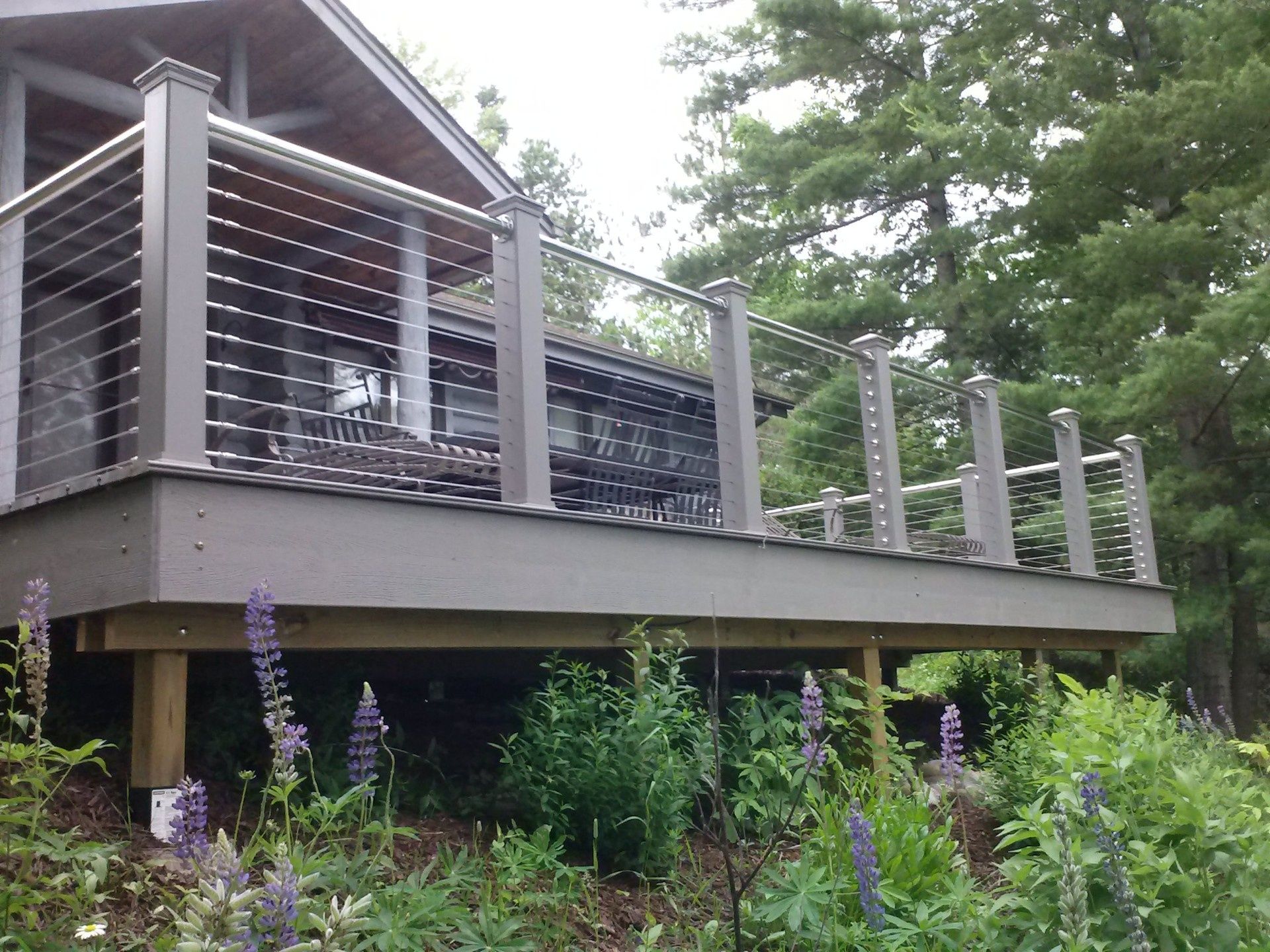 Wooden deck with metal railings on a house, surrounded by lush green trees and purple flowers.