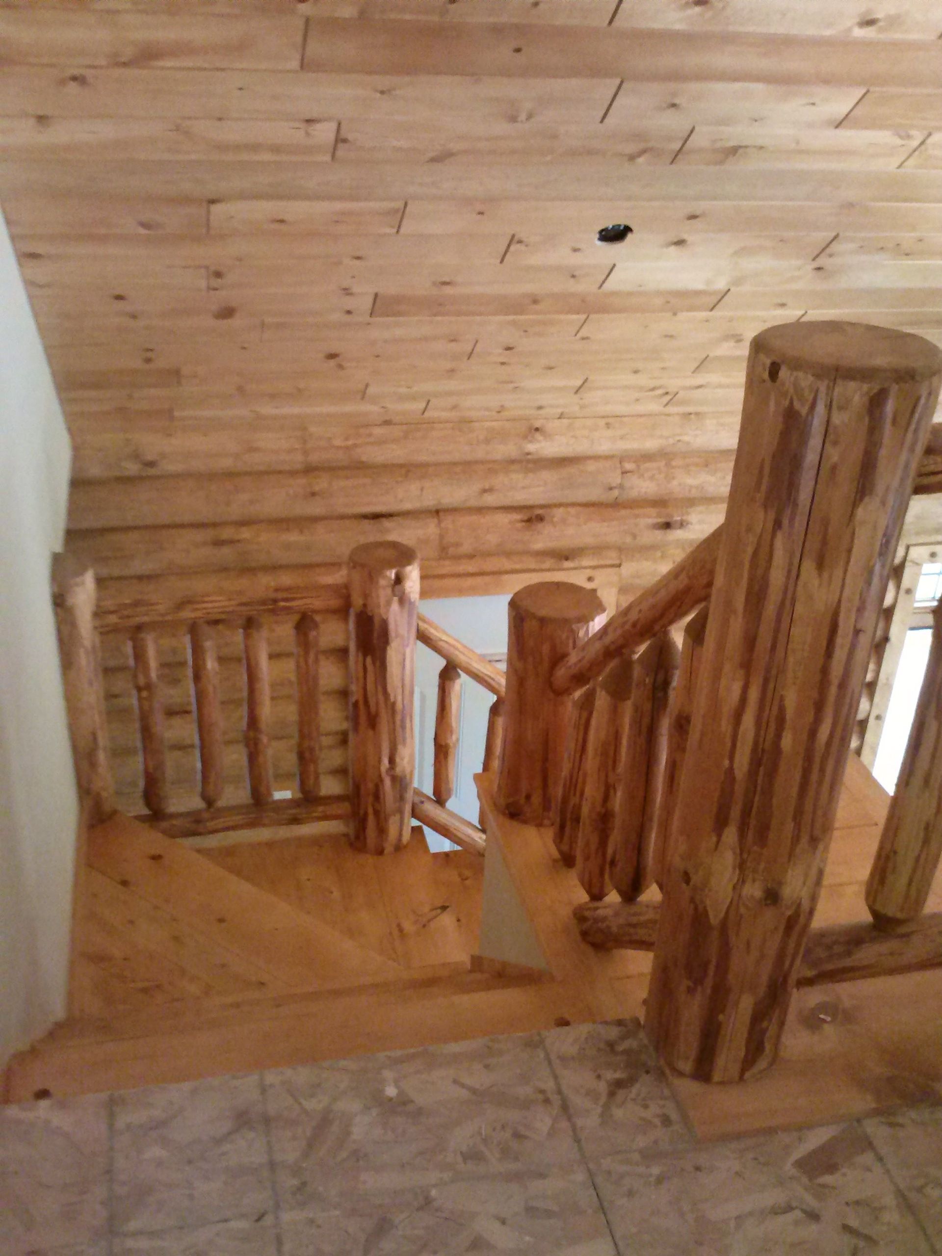Wooden indoor staircase with log railings descending to a lower floor beside a white wall.