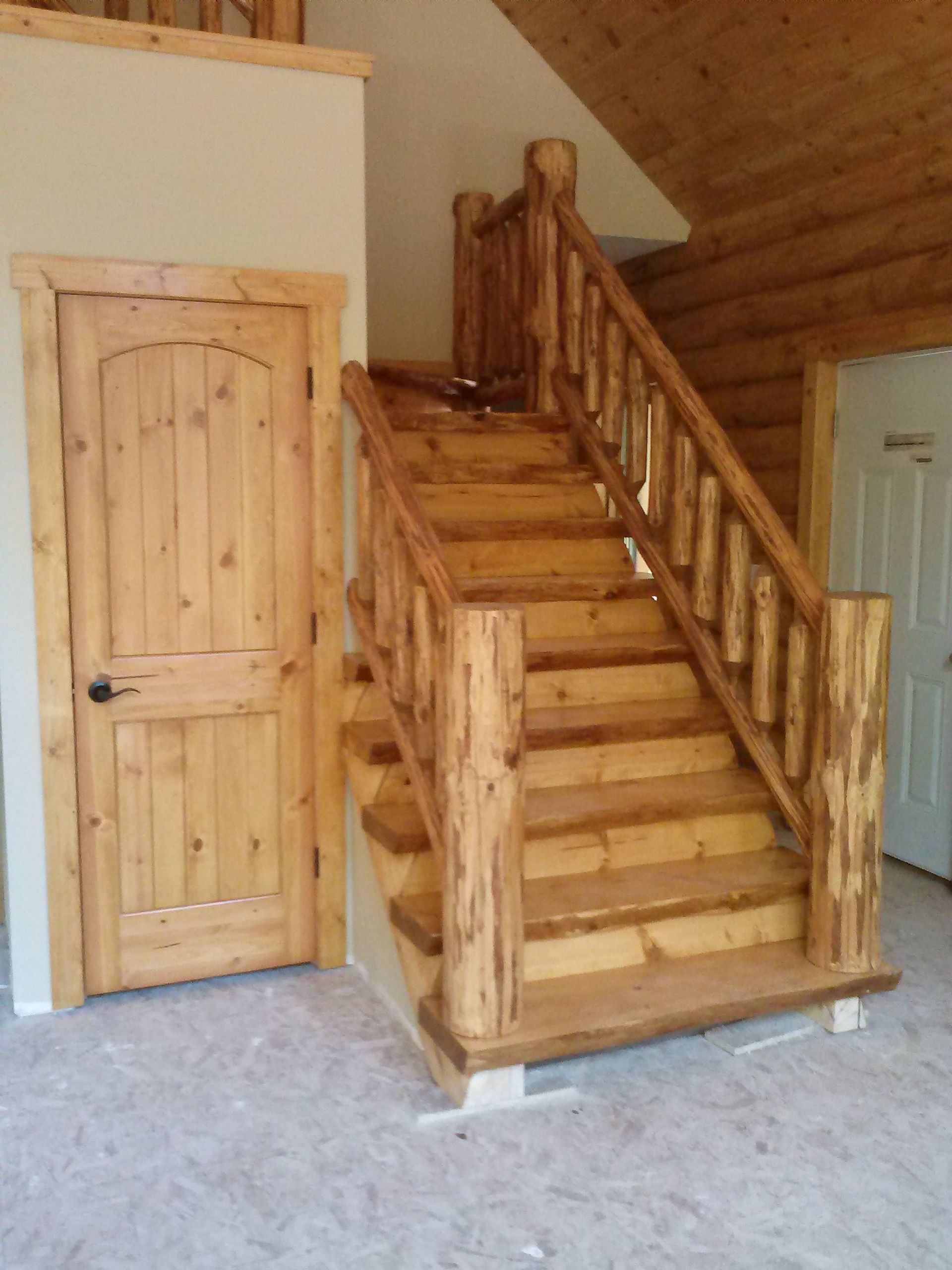 Log cabin staircase with rustic wood railing beside a wooden door on carpeted floor