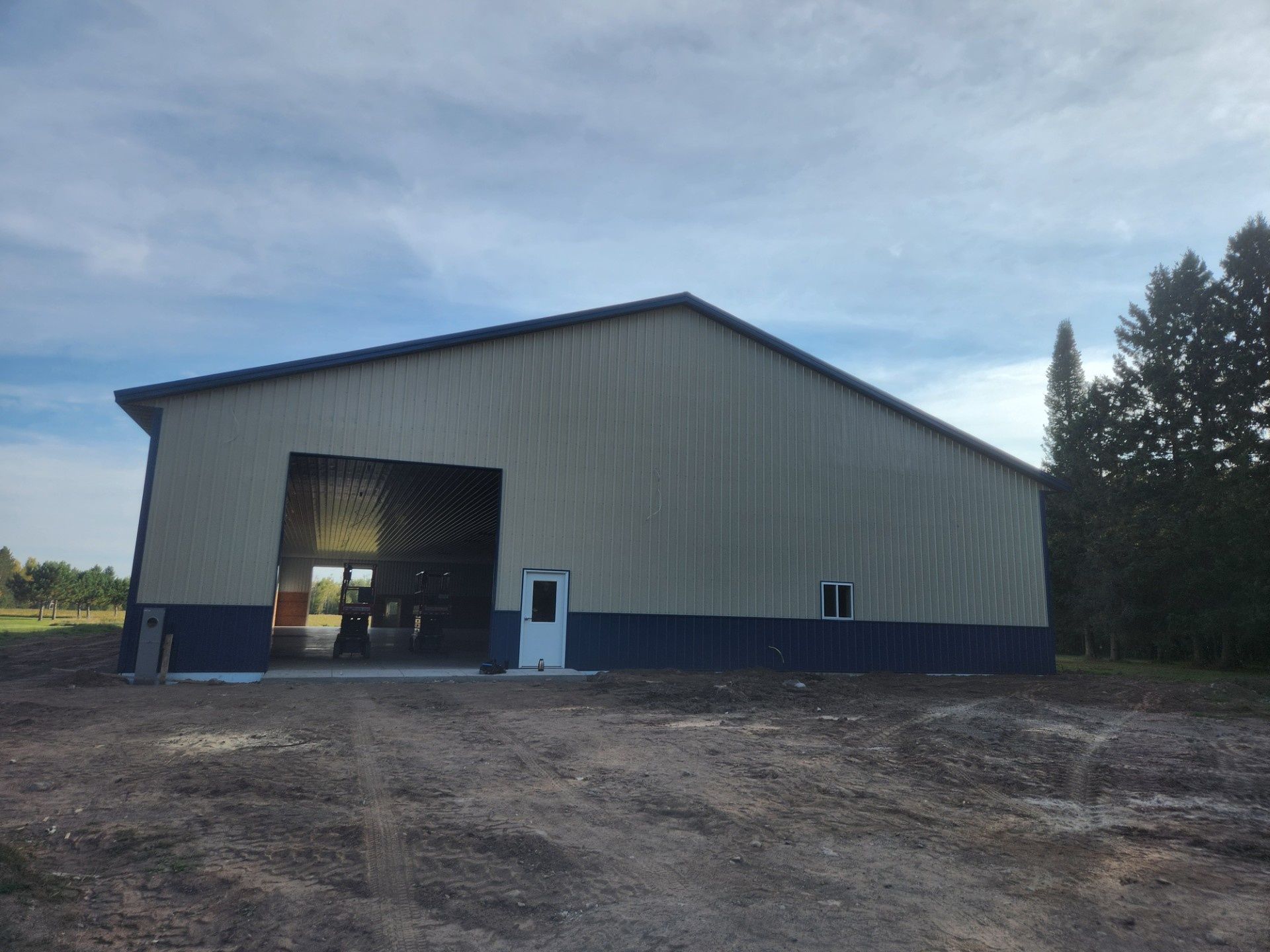 Large gray metal barn with an open bay door on a dirt lot under a cloudy sky