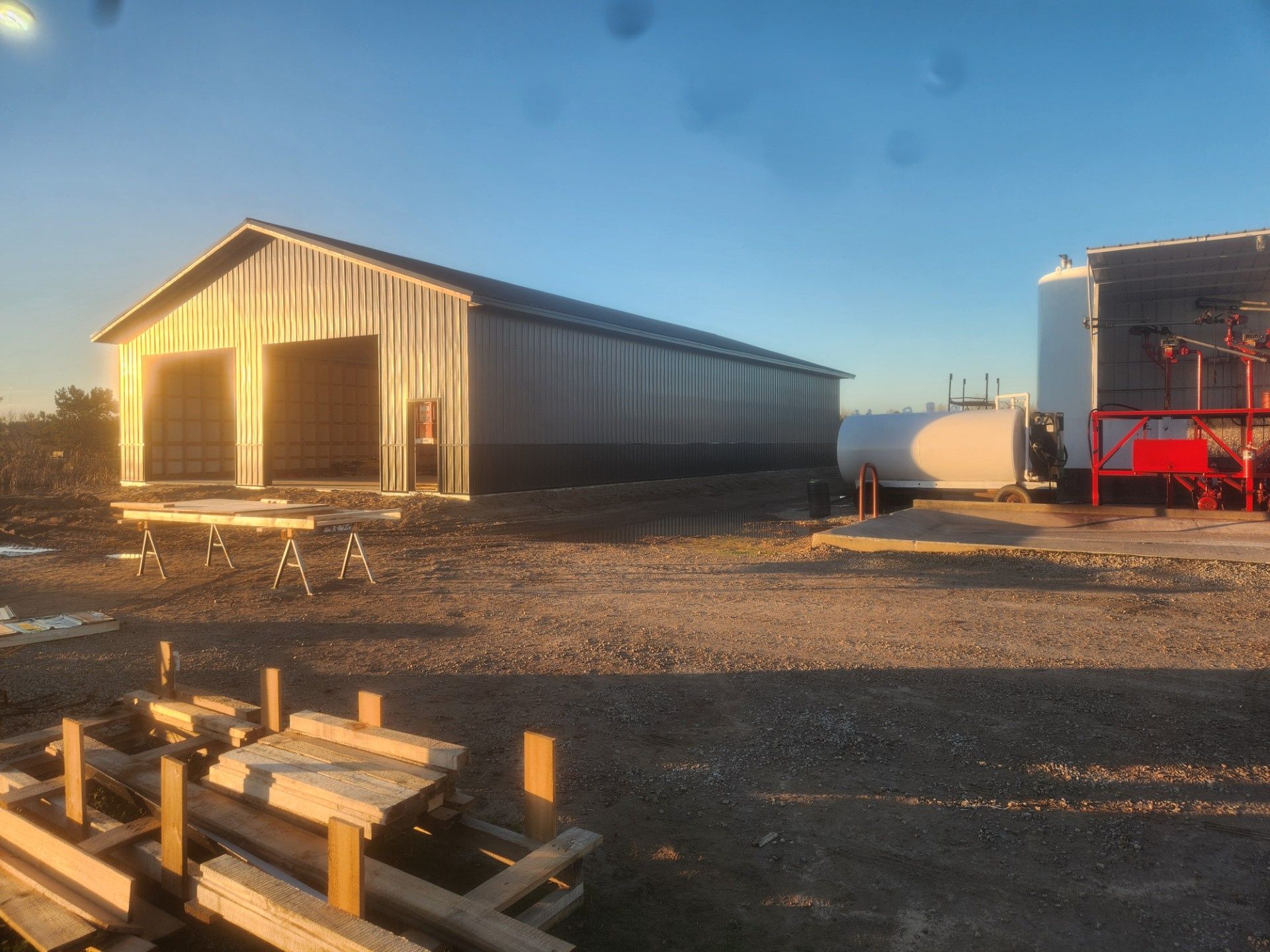 Sunlit industrial yard with metal buildings, tanks, and stacked lumber on gravel.