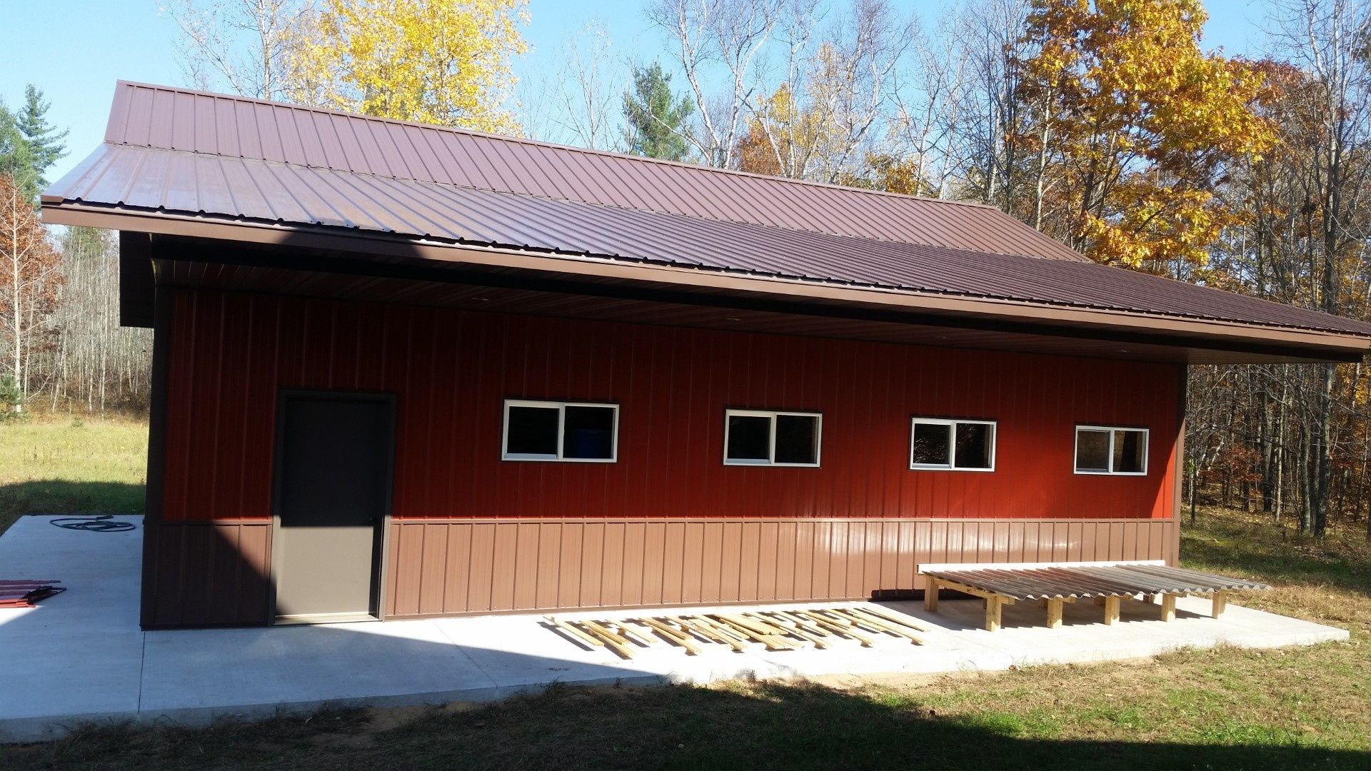 Red barn-like building with a dark roof, small windows, and a bench in front, surrounded by autumn trees.