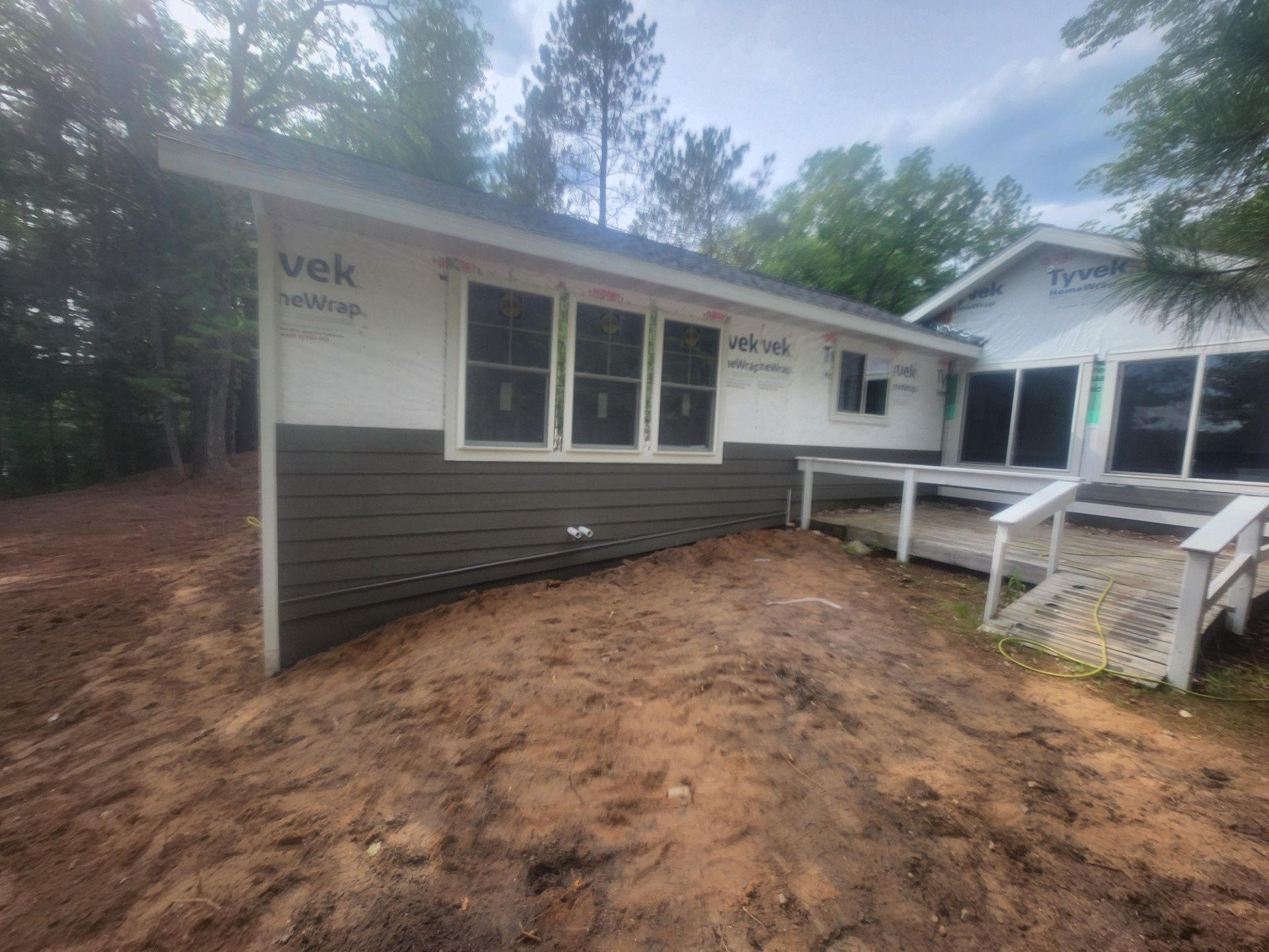 Small white house with dark gray siding, a porch, and muddy ground surrounded by trees