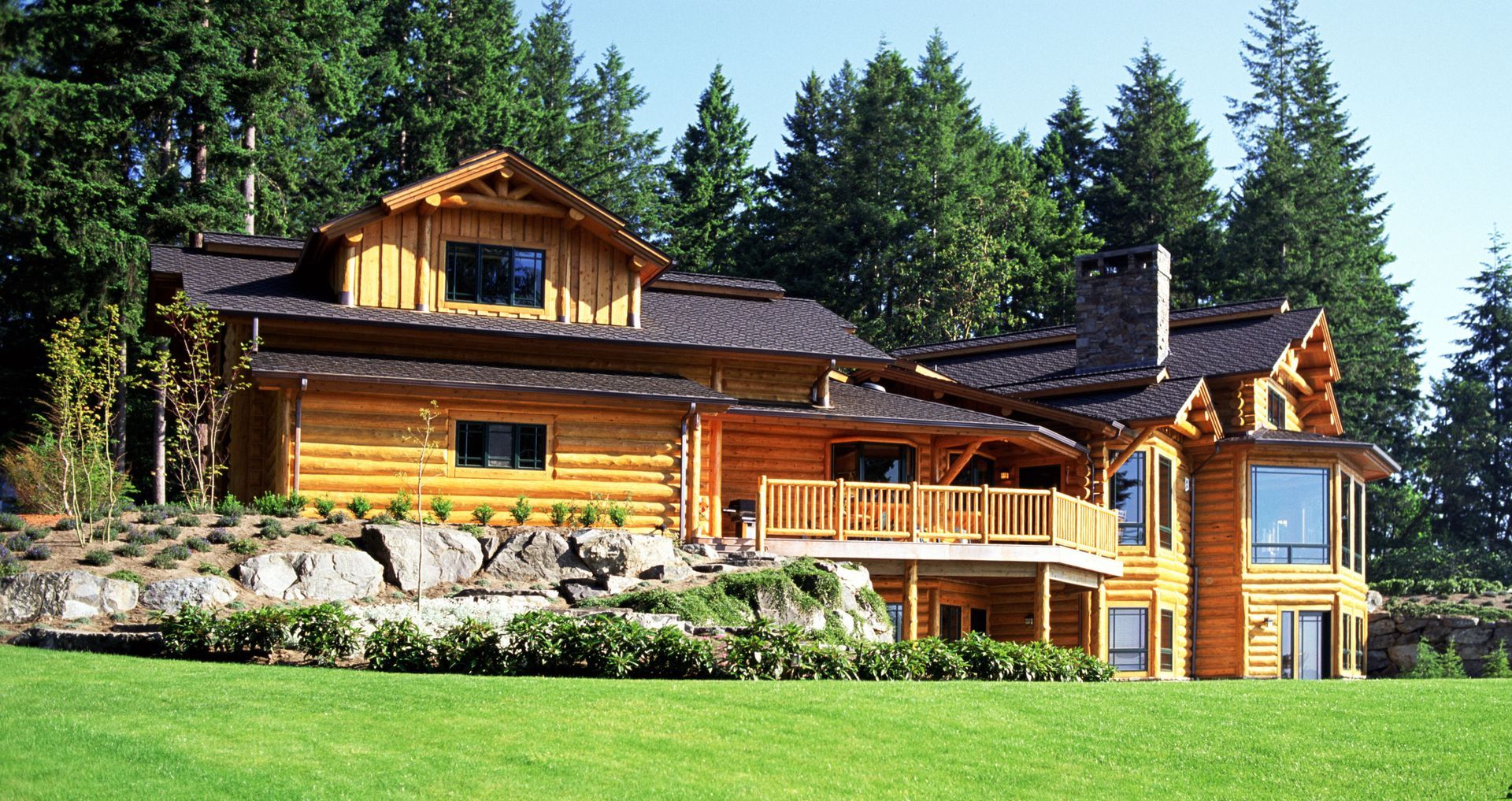 Log cabin house with log siding, stone accents, and a green lawn, surrounded by tall pine trees.
