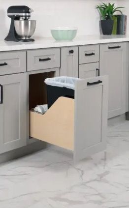 Gray kitchen cabinet with pull-out trash can. A white countertop, appliances and plants sit behind the cabinet.
