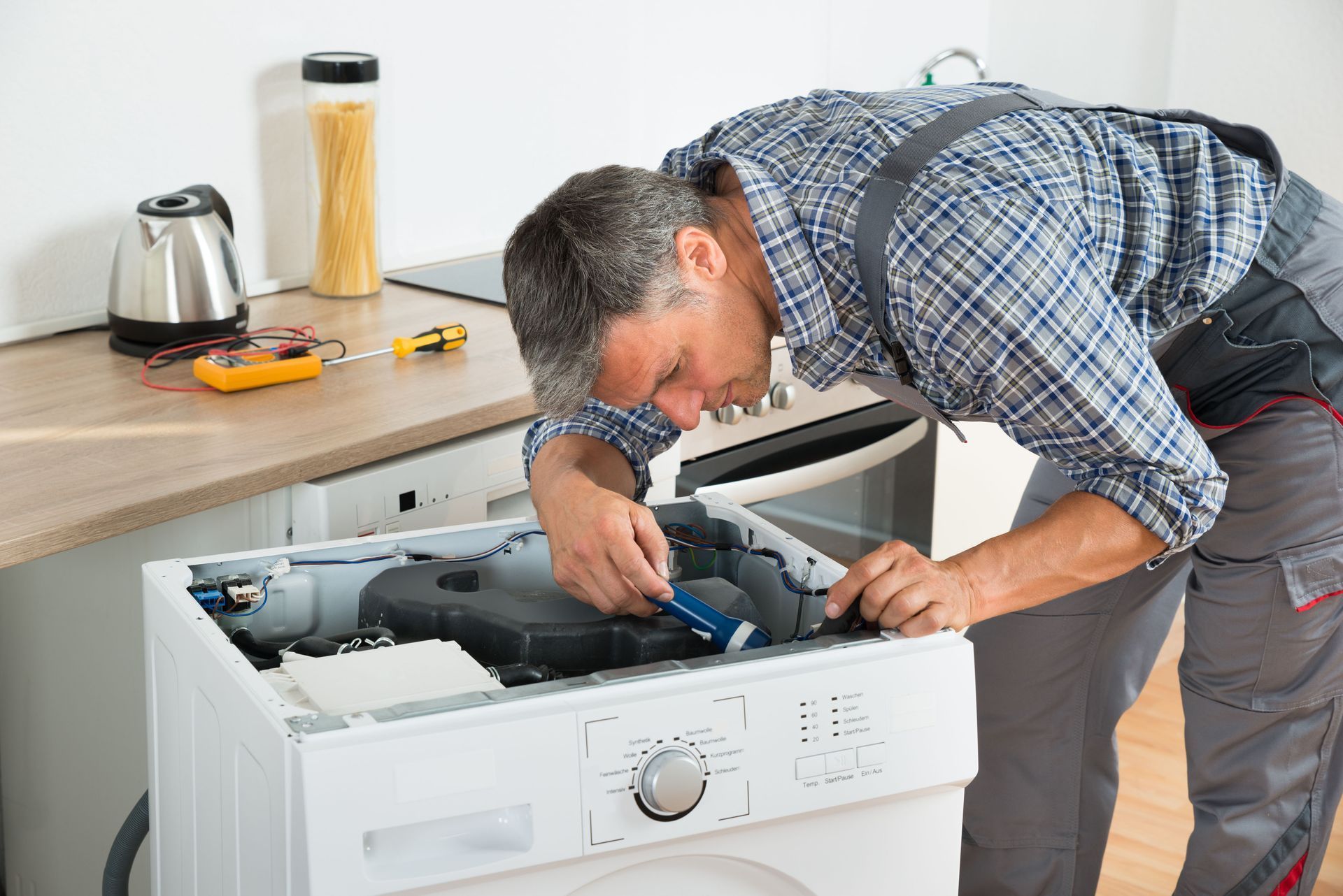 Technician repairing a washing machine in a kitchen, with tools on the counter.