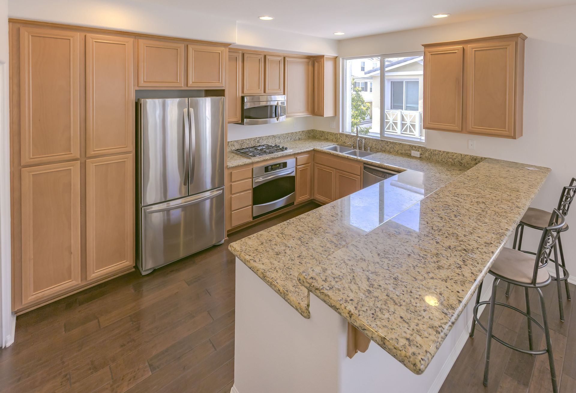 Bright modern kitchen with granite island, stainless steel appliances, wood cabinets, and bar stools